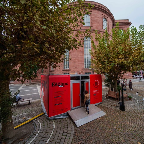 Das Bild zeigt einen rot verkleideten Container auf dem Paulsplatz in Frankfurt. Es handelt sich um einen Escape Room zur Aktion "The Great Escape Room – Eine inspirierende Auszeit auf den Spuren Goethes Italienischer Reise". Im Hintergrund ist die Paulskirche aus rotem Sandstein zu sehen. 