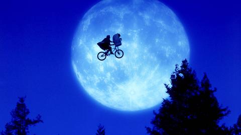 Ein Mensch auf einem Rad mit Fahrradkorb in der Luft über dunklen Bäumen vor einem hellen Mond in blauem Himmel.