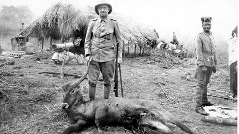 Eine historische Aufnahme zeigt einen europäischen Jäger in Safari-Kleidung. Er steht neben einem erlegten Antilopen-artigen Tier mit großen Hörnern. Im Hintergrund befinden sich einfache Hütten mit Strohdächern. Daneben steht ein weiterer Mann in Uniform. Weitere Personen sind im Hintergrund zu sehen.