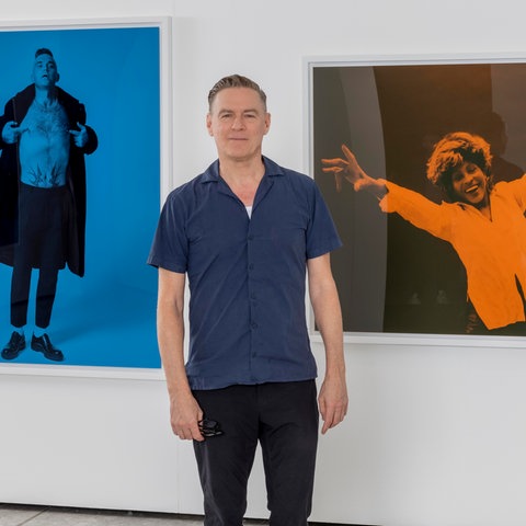 Ein Mann steht vor einer Wand, an der drei Bilder hängen.