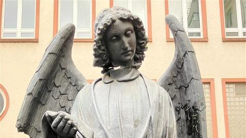 Eine Steinskulptur eines Engels steht vor einem Gebäude mit Fenstern. Der Engel hat lockiges Haar, trägt ein langes Gewand und hält eine Schriftrolle in den Händen. Ein Flügel ist gebrochen. Im Hintergrund sind Büsche und die Fassade des Gebäudes zu sehen.