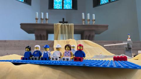 Das Bild zeigt mehrere sitzende Lego-Männchen, die auf eine blaue Platte geklemmt sind. Im Hintergrund ist ein Altar mit Kreuzen zu sehen.