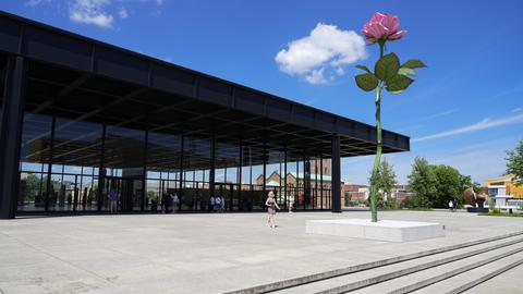 Ein modernes Gebäude mit einer Glasfassade und dunklem Dach, im Vordergrund eine große Skulptur einer pinkfarbenen Rose auf einem hohen Stiel. Vor dem Gebäude gehen Menschen über eine gepflasterte Fläche. Der Himmel ist blau mit wenigen Wolken.