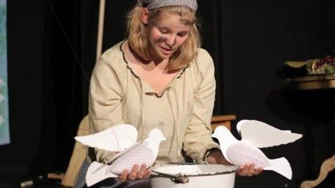 Eine junge Frau mit blonden Haaren sitzt auf einer Bühne. Sie ist in Fetzen gehüllt, in der Hand hat sie zwei Tauben aus Pappe. Es handelt sich um eine Schauspielerin, die am Gießener Theater "Aschenputtel" darstellt. 