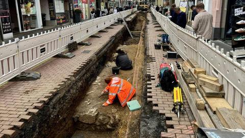 Das Bild zeigt eine in der Mitte aufgerissene Fußgängerzone. Zwei Bauarbeiter sitzen in dem freigelegten Bereich und graben Teile einer alten Mauer aus. Links und rechts gehen Menschen an Geschäften vorbei.