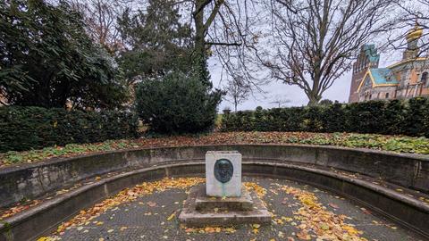 Leerer Sockel auf einem Platz, dahinter eine Hecke, Bäume und rechts ein Gebäude mit teils goldenem Dach.