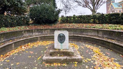 Leerer Sockel auf einem Platz, dahinter eine Hecke, Bäume und rechts ein Gebäude mit teils goldenem Dach.