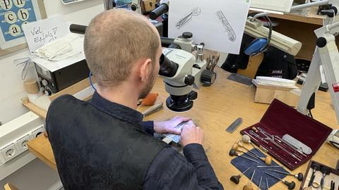 Ein Mann sitzt an einem Tisch und blickt durch ein Mikroskop. Seine Hände arbeiten an einem kleinen Stück, viele kleine Werkzeuge liegen auf dem Tisch. Eine Skizze hängt vor ihm.