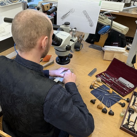 Ein Mann sitzt an einem Tisch und blickt durch ein Mikroskop. Seine Hände arbeiten an einem kleinen Stück, viele kleine Werkzeuge liegen auf dem Tisch. Eine Skizze hängt vor ihm.