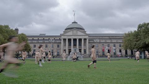 Nackte Menschen laufen über eine Wiese, im Hintergrund das klassizistische Gebäude des Staatstheaters Wiesbaden