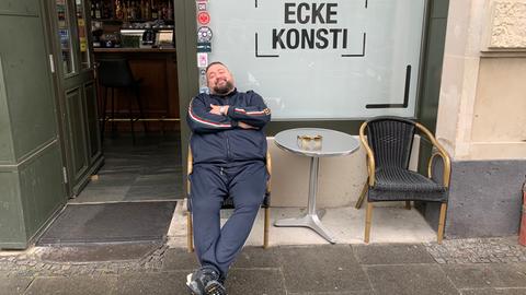 Ein Mann im Trainingsanzug sitzt lässig in einem Sessel vor einem Café.