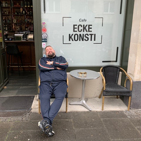 Ein Mann im Trainingsanzug sitzt lässig in einem Sessel vor einem Café.