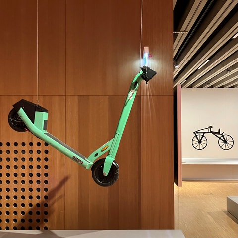 Das Bild zeigt einen Ausstellungsraum im Historischen Museum Frankfurt. Zu sehen ist ein türkisfarbener E-Scooter, der von einer holzvertäfelten Decke hängt.