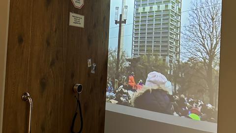 Das Bild zeigt eine Ausstellungsansicht von "Zeitzeugen? Ein Erinnerungslabor" im Historischen Museum Frankfurt. Zu sehen ist ein nachgebauter Paternoster, im Hintergrund läuft eine Foto-Show auf einer Leinwand.