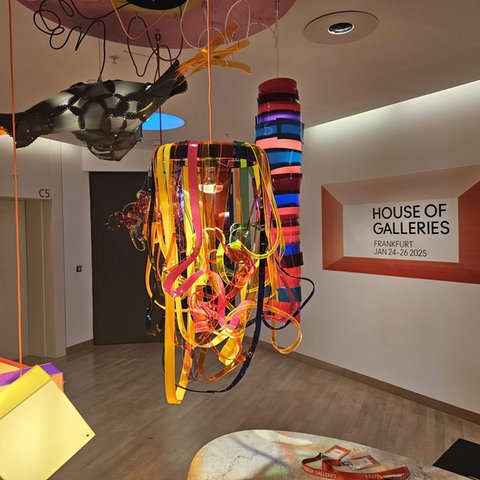 Eine moderne Kunstinstallation mit bunten, geschwungenen Elementen hängt von der Decke. Im Hintergrund befindet sich ein Schild mit der Aufschrift "HOUSE OF GALLERIES, FRANKFURT, JAN 24-26 2025". Links ist ein geschlossener Aufzug sichtbar. Auf einem Tisch darunter liegt ein rotes Band. Der Raum hat helle Holzböden und weiße Wände.