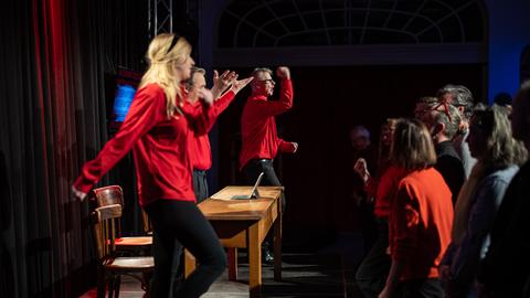 Eine Gruppe von Personen in roten Hemden steht auf einer Bühne und bewegt sich rhythmisch. Vor ihnen steht ein Holztisch mit einem kleinen Gerät darauf. Im Hintergrund sind Vorhänge und schwaches Licht zu sehen. Zuschauer in verschiedenen Kleidungsstücken, darunter ein rotes Oberteil, stehen vor der Bühne.