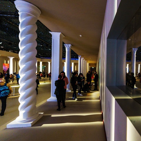 Das Bild zeigt den Ehrengast-Pavillon von Buchmesse-Gastland Italien. Zu sehen ist eine spärlich beleuchtete Halle mit weißen Säulen. Dazwischen stehen Menschen, die sich unterhalten.