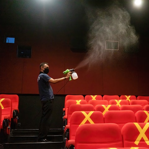 Ein Mann mit schwarzer Maske steht in einem Kino und desinfiziert mit einem Sprühgerät. Die roten Kinositze sind teilweise mit gelbem Klebeband in X-Form markiert. Es ist wenig Licht im Raum.