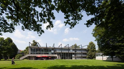 Ein langgezogenes Gebäude mit einer großen Terrasse. Es ist Sommer, im Vordergrund ist eine Wiese.