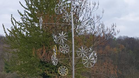 Die Skulptur "FahrRat" aus Edelstahl und Aluminium zeigt mehrere Fahrradreifen, die wie Felgen anmuten. Sie sind an einer Art Laternenpfahl montiert.