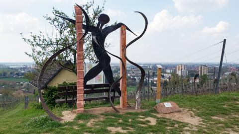 Das Bild zeigt eine Skulptur inmitten von Weinbergen. Sie ist aus Sandstein und Eisen gefertigt und zeigt angedeutet einen Menschen in Bewegung.