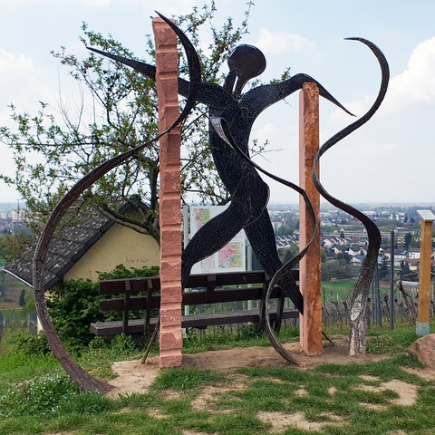 Das Bild zeigt eine Skulptur inmitten von Weinbergen. Sie ist aus Sandstein und Eisen gefertigt und zeigt angedeutet einen Menschen in Bewegung.