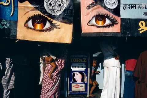 Blick auf ein Geschäft, über dem Plakate mit großen Augen hängen. Darunter stehen Frauen in weißen Saris. Ihre Köpfe sind von den Augen-Plakaten bedeckt.