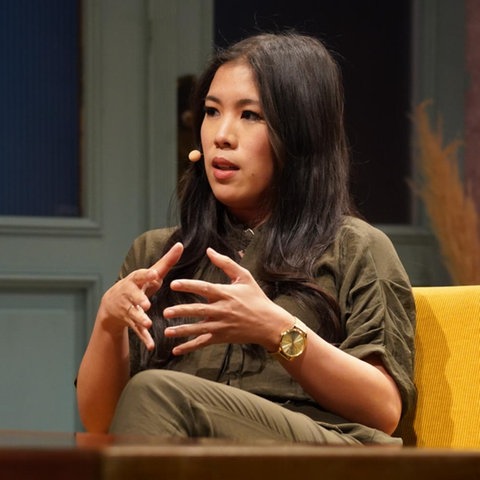 Mai Thi Nguyen-Kim sitzt auf einem gelben Sofa. Sie trägt einen khakifarbenen Overall und ihre dunklen Haare fallen über ihre Schulter.