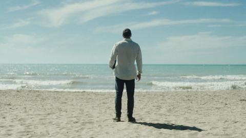 Das Bild zeigt einen hellen Sandstrand und das türkisblaue Meer dahinter. Am Strand steht ein Mann in Jeans und hellem Hemd. Er blickt auf das Meer und ist von hinten zu sehen. Der Himmel ist blau und von zarten Wolken überzogen.