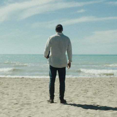 Das Bild zeigt einen hellen Sandstrand und das türkisblaue Meer dahinter. Am Strand steht ein Mann in Jeans und hellem Hemd. Er blickt auf das Meer und ist von hinten zu sehen. Der Himmel ist blau und von zarten Wolken überzogen.