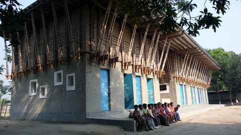 Das Bild zeigt ein zweistöckiges Schulgebäude in Bangaldesch, vor dem zwölf Kinder in einer Reihe sitzen. Das Gebäude besteht aus einem aus Lehm gefertigen grauen rechteckigen Bau mit einer langgezogenen Treppe davor. Das Dach besteht aus Bambusstangen, die zu einer Art Zelt aufgestellt sind.