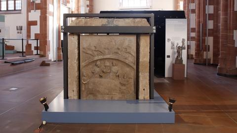 Das Bild zeigt das Archälogische Museum Frankfurt im ehemaligen Karmeliterkloster. Zu sehen ist eine Kirche mit hohen Decken sowie Pfeilern aus rotem Sandstein und weißen Kacheln. In der Mitte des Raumes ist ein Kultbild der römischen Gottheit Mithras aus der Römerstadt Nida zu sehen: ein großer rötlicher Sandstein, in den Figuren geschlagen wurden. Der Stein wird gestützt von dunkelgrauen Pfeilern und ist in einen hellblau gestrichenen erhöhten Fußboden eingelassen.