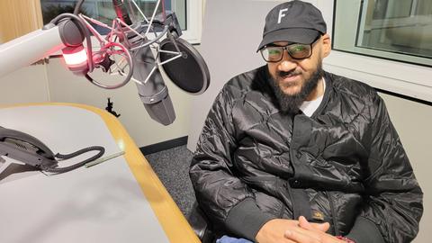 Das Bild zeigt Rapper Moses Pelham in einem hr-Studio. Zu sehen ist ein Mann mit schwarzer glänzender Bomberjacke, einer schwarzen Basecap und einer schwarz gerahmten Brille. Er trägt einen dunklen Ziegenbart und sitzt auf einem Stuhl. Am linken Bildrand sind ein Mikrofon und ein Tisch zu sehen.