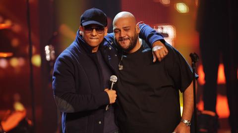 Das Bild zeigt den Rapper Moses Pelham und Sänger Xavier Naidoo. Zu sehen sind ein Mann mit dunkler Schiebermütze und Sonnenbrille, der einen anderen Mann mit Glatze und schwarzem T-Shirt umarmt. Beide halten ein Mikrofon in der Hand.