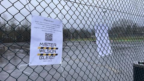 An einem Drahtzaun hängt ein laminiertes Schild. Darauf steht "Mister Wilson bleibt". Es macht Werbung für die Online-Petition gegen den Teilabriss. Darauf ist ein QR-Code gedruckt, der direkt zur Petition führt. Im Hintergrund ist ein Asphaltplatz mit Skater-Rampen.