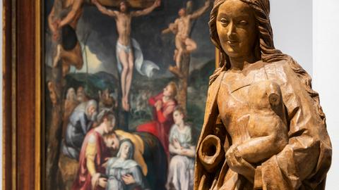 Im Vordergrund eine Holzskulptur der Jungfrau Maria, im Hintergrund ein großformatiges Gemälde, das die Kreuzigung Jesu zeigt.