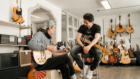 Das Bild zeigt zwei Männer in einem Musikladen. Ein älterer Mann mit langen Haaren und einem Bandana sitzt auf einem sogenannten Cajon, einem Holzkasten zum Trommeln. Er hält eine beigefarbene Gitarre in den Händen und blickt auf seine Finger auf den Saiten. Neben ihm sitzt ein jüngerer Mann mit schwarzen Haare und schwarzem T-Shirt auf einem Barhocker. Er hat ebenfall seine Gitarre auf dem Schoß. Seine Hände liegen vor dem Instrument auf der Sitzfläche des Stuhls. Im Hintergrund sind weitere Gitarren an einer Wand zu sehen. 