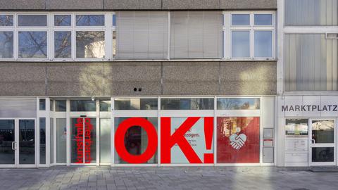 Das Bild zeigt die Fassade eines Gebäudes mit großen Fenstern. Auf den Glastüren links steht "Kunsthalle Offenbach" in roter Schrift. Rechts sind Fenster mit einer großen roten Aufschrift: "OK!" und einem Symbol mit zwei sich schüttelnden Händen. Daneben steht "MARKTPLATZ 1" auf einer weißen Wand.