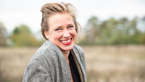 Eine lächelnde Frau mit rotem Lippenstift trägt eine graue Jacke. Sie steht im Freien vor einem unscharfen, naturbelassenen Hintergrund mit Bäumen und Wiese.