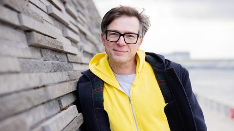 Ein Mann mit Brille und leuchtend gelber Jacke lehnt an einer Wand.