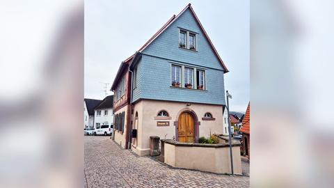 Ein zweistöckiges Haus mit spitzem Dach. Das obere Stockwerk ist blau, unten hellbraun. Unten ist die Tür, im ersten Stock drei Fenster, im Dachgeschoss zwei. Davor steht eine Mauer.