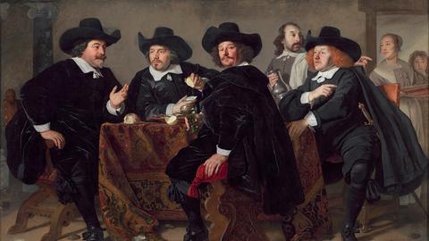 Das Bild zeigt das Gemälde "Die Vorsteher des Kloverniersdoelen" des Künstlers Bartholomeus van der Helst aus dem Jahr 1655. Zu sehen sind vier Männer mit schwarzen Umhängen, weißen Kragen und schwarzen Hüten. Sie sitzen breitbeinig um einen Tisch herum. Im Hintergrund sind ein weiterer, einfacherer gekleideter Mann und eine Frau zu sehen.