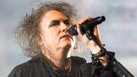 Das Bild zeigt den Musiker Robert Smith, Frontmann der Gothic-Band The Cure. Er trägt sein langes graues Haar wild zerzaust und Eyeliner um die Augen.