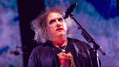 Das Bild zeigt Musiker Robert Smith, Frontmann der Gothic-Band The Cure. Er trägt seine grauen langen Haare wild auftoupiert und Eyeliner um seine Augen.