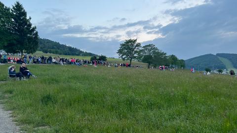 Menschen sitzen außerhalb des Konzertgeländes zum Teil mit Campinstühlen auf einer Wiese und genießen die Musik.