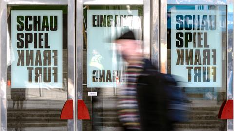 Ein Mann läuft vor gläsernen Türen entlang. An den Türen hängen Plakate mit der Aufschrift "Schauspiel Frankfurt".