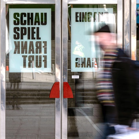 Ein Mann läuft vor gläsernen Türen entlang. An den Türen hängen Plakate mit der Aufschrift "Schauspiel Frankfurt".