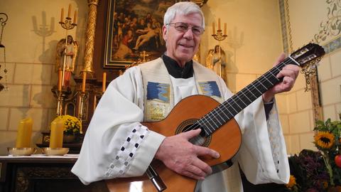 Ein Pfarrer steht mit einer Gitarre in einer Kirche und singt v