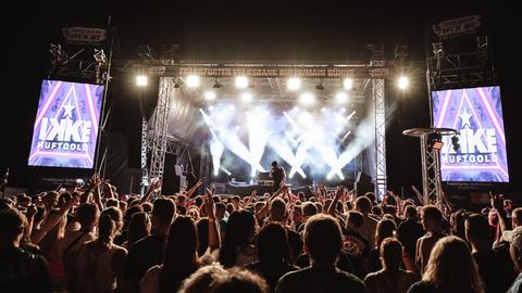 Das Bild zeigt das Musikfestvial Sossenheim Open Air. Zu sehen ist eine beleuchtete Bühne mit Publikum im Vordergrund. Auf Bannern neben der Bühne steht "Ikke Hüftgold".