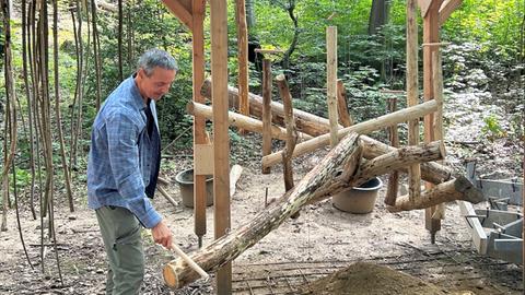 Das Bild zeigt Künstler Volker Staub. Er steht vor einer Installation aus Holzstämmen und Metallrohren. Mit einem Stöckchen bringt er das wie ein Windspiel angeordnete Kunstwerk zum Klingen.